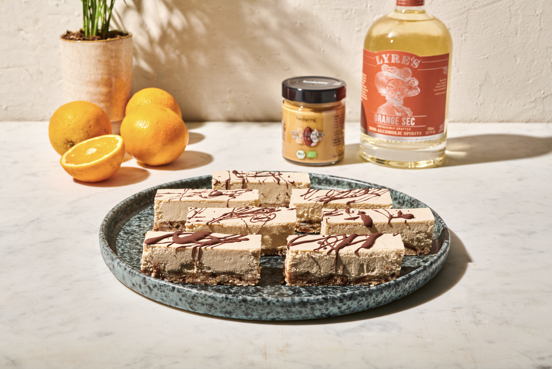 Plate of cheesecake bars with chocolate drizzle on a white table, with oranges, a potted plant, a jar of peanut butter, and a bottle of liquor in the background.