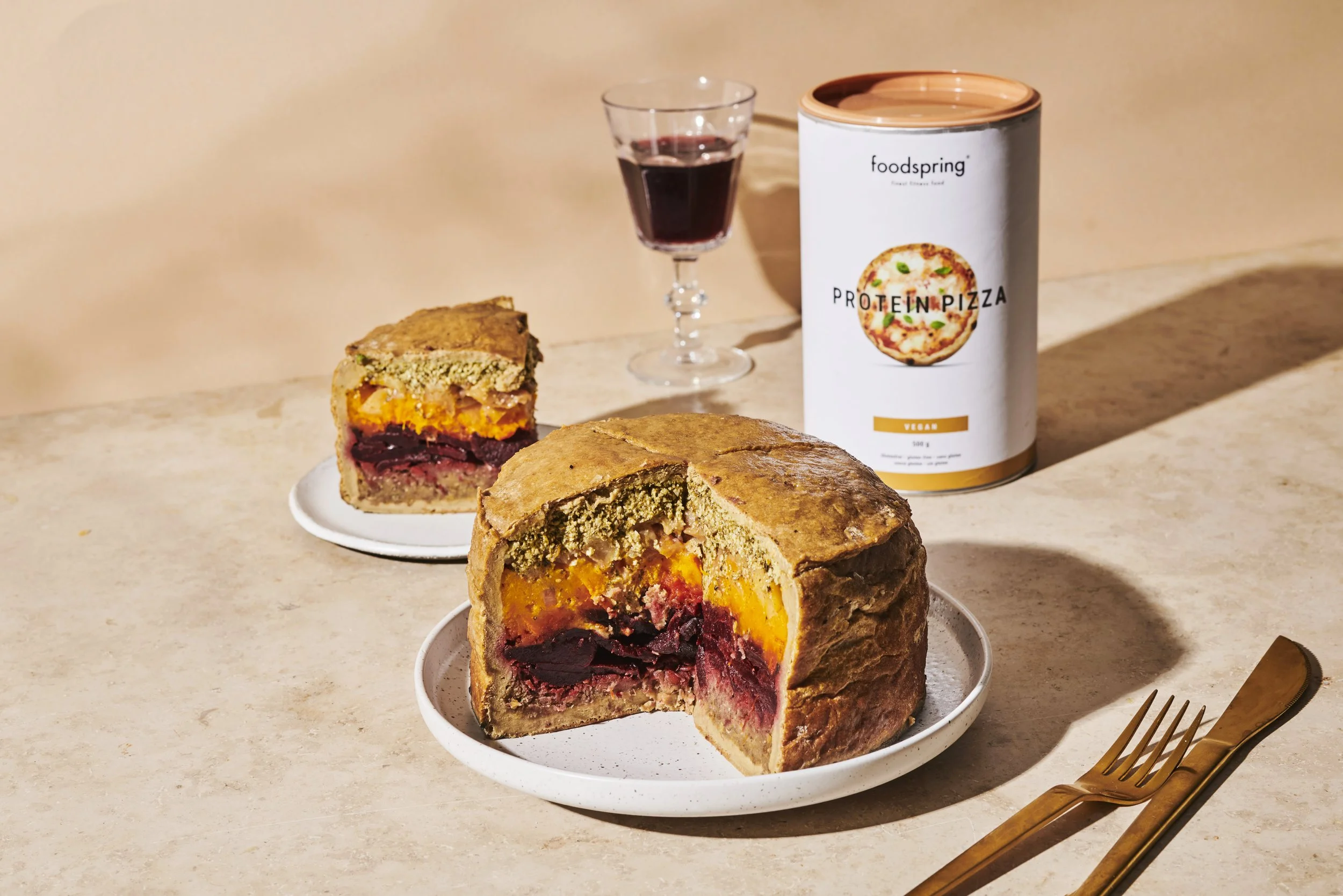 Whole and sliced vegan protein pizza with a side of red wine and a can of Foodspring protein pizza mix on a beige surface.