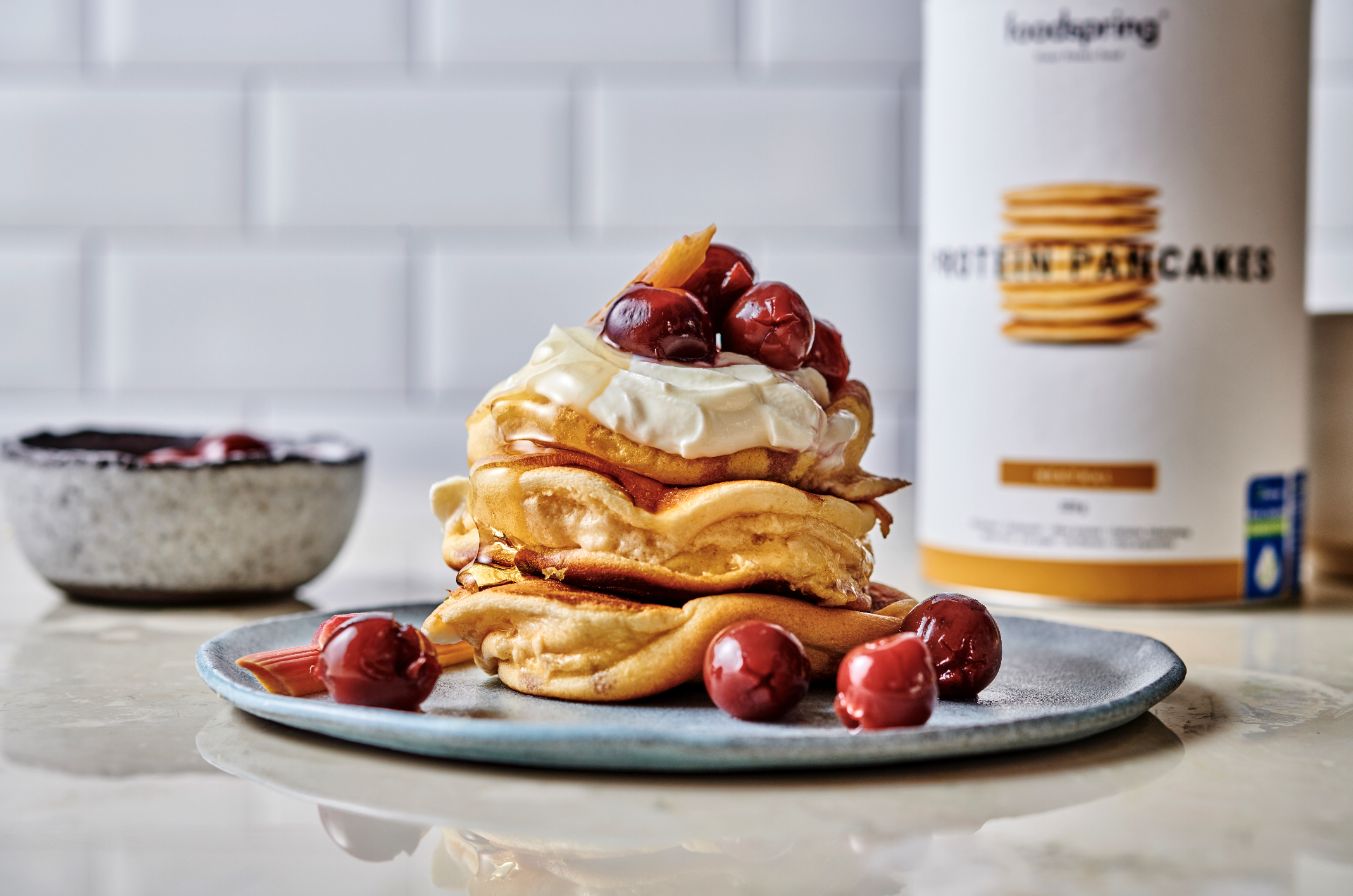 Stack of pancakes topped with whipped cream and cherries, served on a gray plate with additional cherries and syrup, with a container of pancake mix in the background.