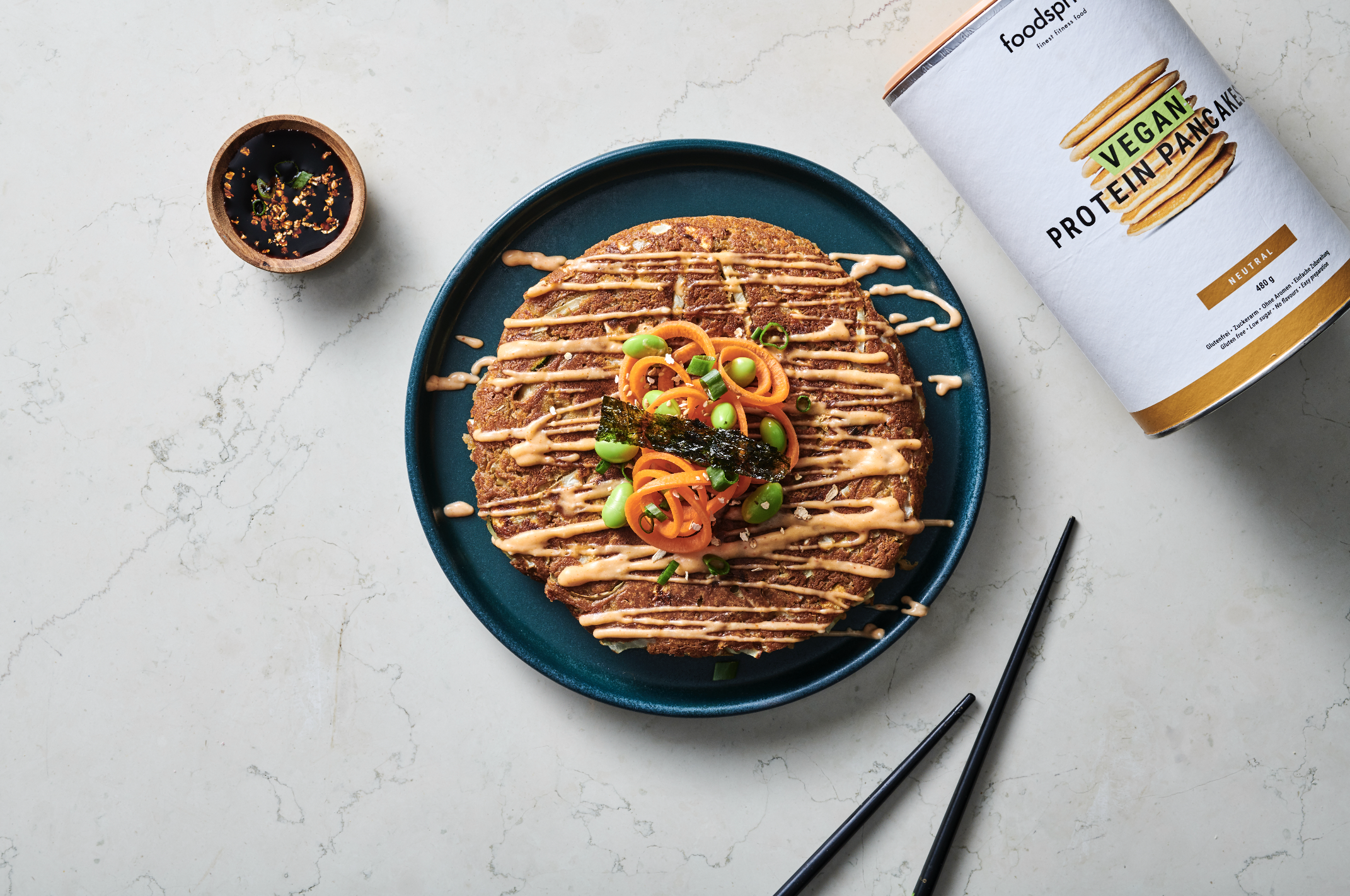Vegan protein pancake topped with chopped scallions, sliced carrots, and a seaweed strip, drizzled with sauce on a teal plate, with soy sauce in a small bowl, chopsticks, and a can of vegan protein pancake mix nearby.