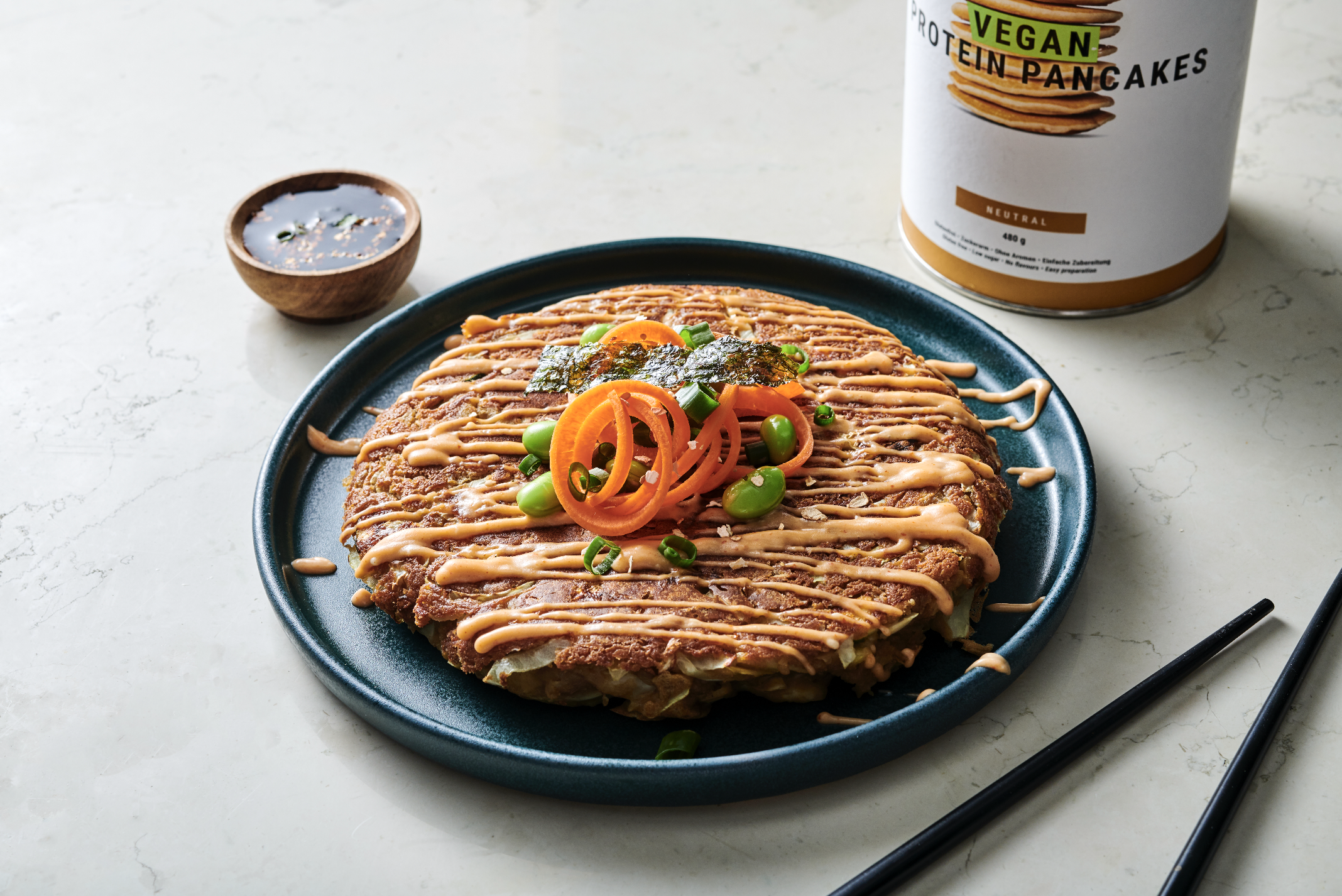Vegan protein pancake topped with sliced carrots, green edamame, and green onions, drizzled with sauce, served on a blue plate with a small bowl of sauce and a container of vegan protein pancake mix in the background.