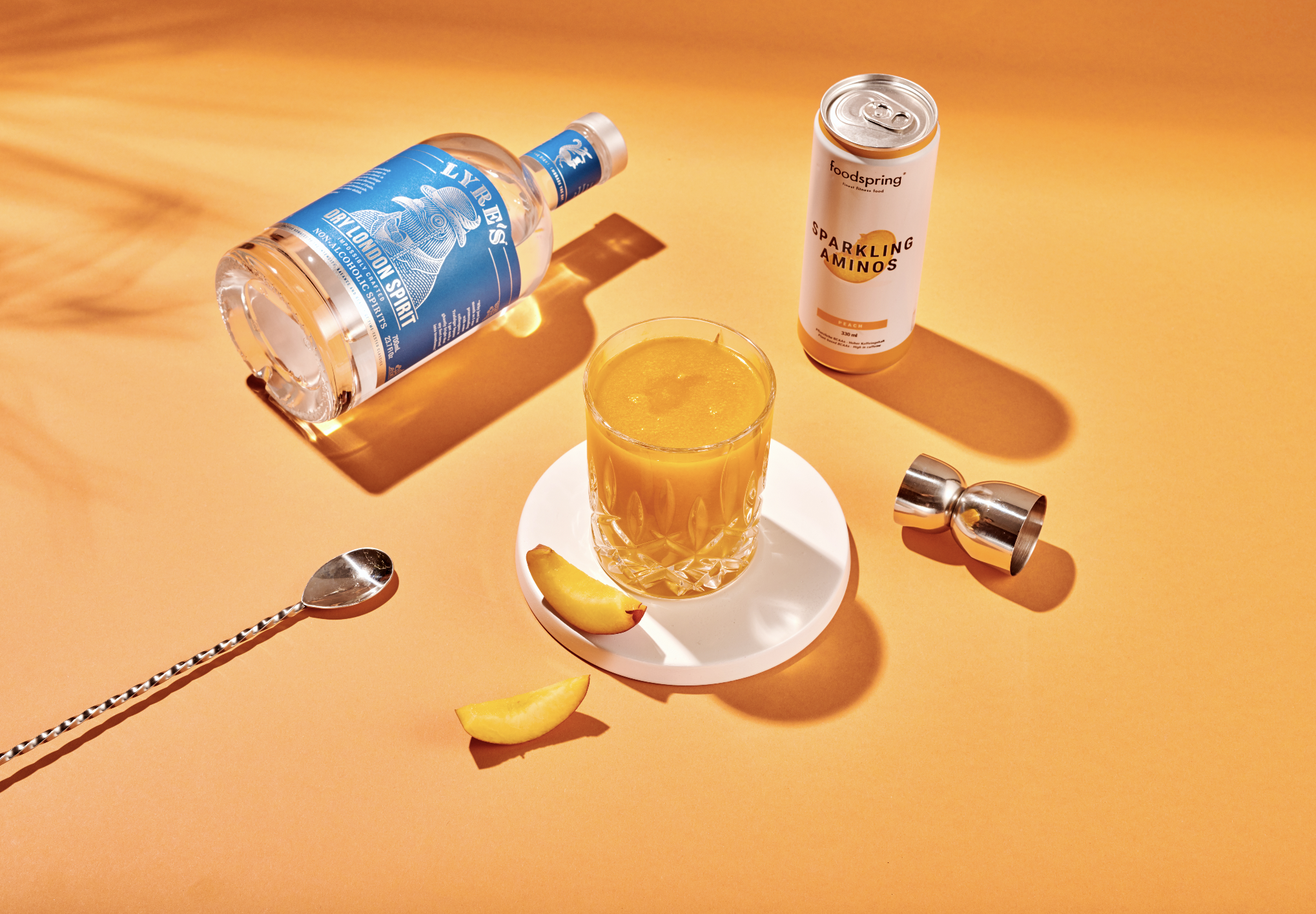 A drink with orange juice in a glass on a white coaster, a peach slice garnish, and a peach wedge on a yellow background with cocktail tools, a bottle of spirits, and a can of sparkling amino drink surrounding it.