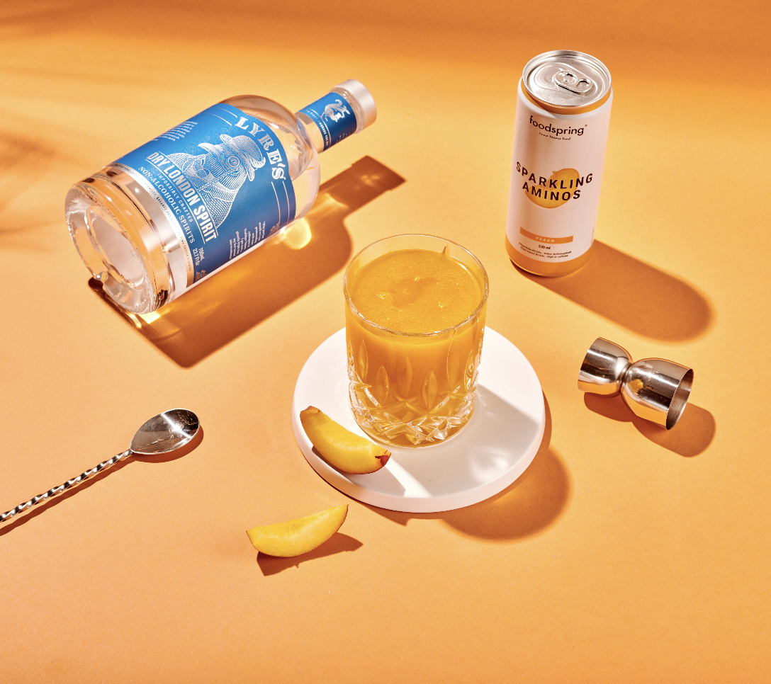 A cocktail glass filled with orange-colored drink on a white coaster, garnished with lemon slices, set on an orange background. Surrounding it are cocktail shakers, a bottle of liquor, a can of sparkling amino drink, a spoon, and lemon wedges.