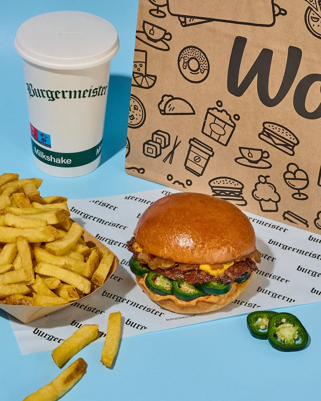 A burger with jalapeños, fries, a milkshake, and a paper bag with food illustrations.