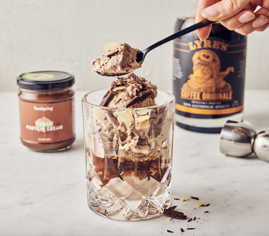 A glass filled with ice, chocolate ice cream, chocolate syrup, and topped with chocolate shavings and sliced almonds, with a spoonful of ice cream being added.