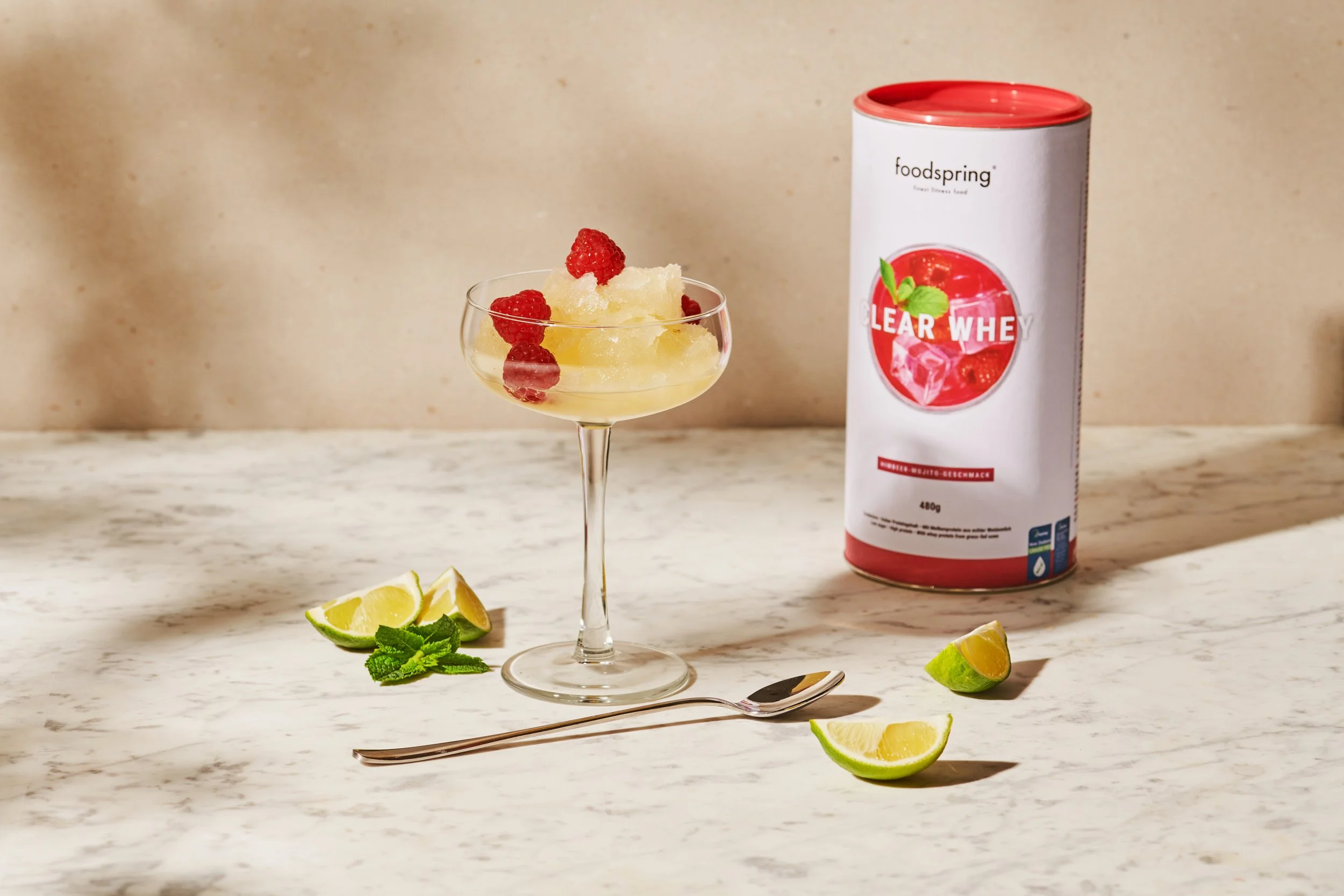 A cocktail glass filled with a yellowish drink garnished with raspberries. Lime wedges and mint leaves on the table, along with a can of clear whey protein powder. A silver spoon is also on the table.