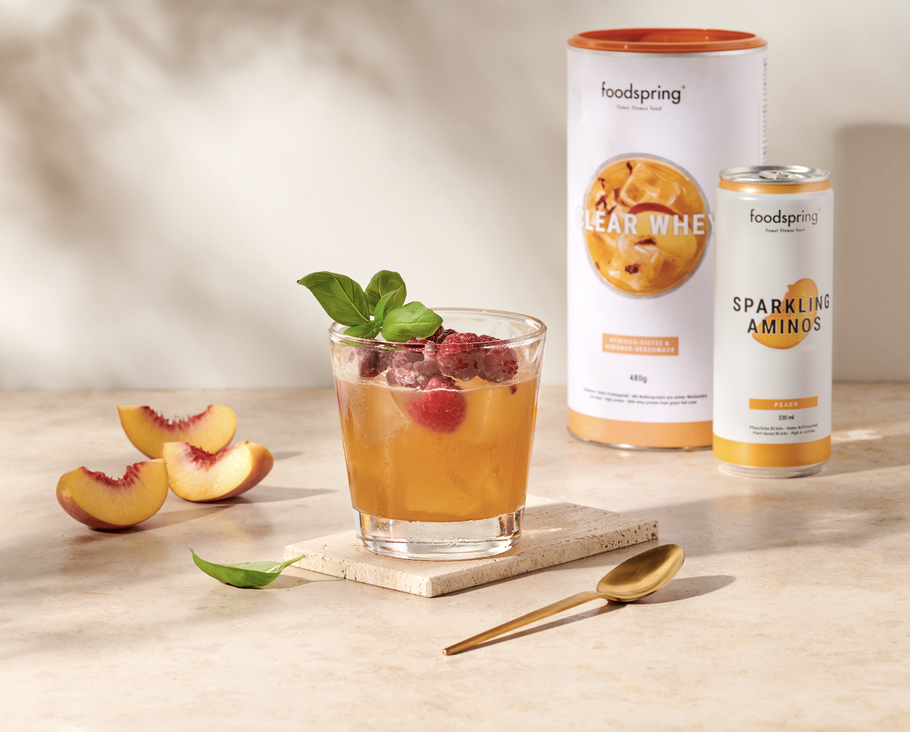A glass of peach-flavored beverage garnished with raspberries and basil leaves, placed on a wooden surface with peach slices beside it, and dietary supplement containers in the background.