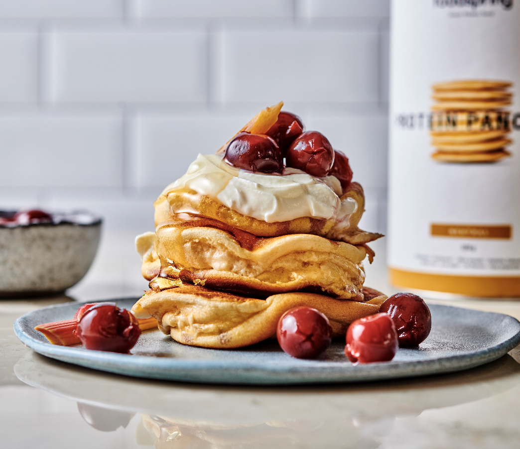 Stack of fluffy pancakes topped with whipped cream, cherries, and a piece of pineapple on a blue plate with additional cherries around it. In the background, there is a container of pancake mix and a bowl of berries.