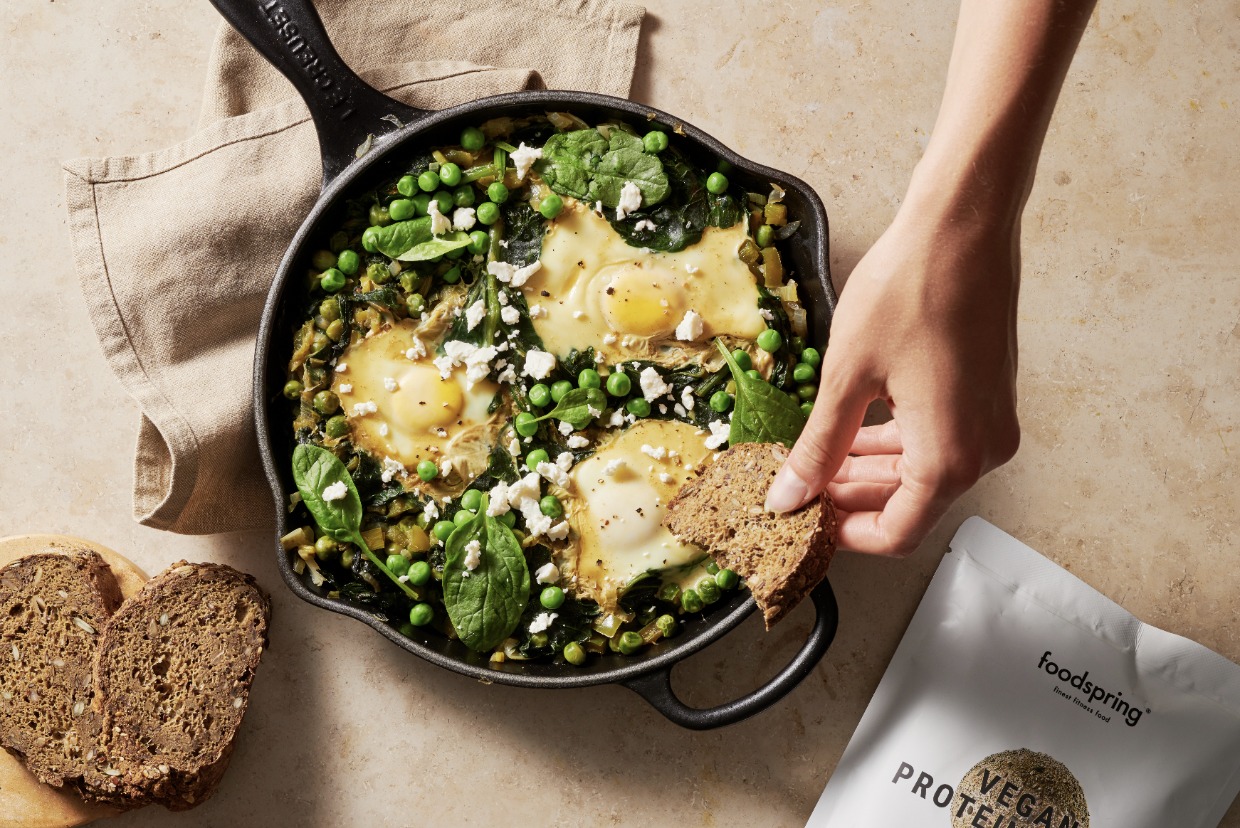 Cast iron skillet with baked eggs, spinach, peas, and crumbled vegan cheese, with a hand holding a slice of bread, and a bag of vegan protein on the counter.
