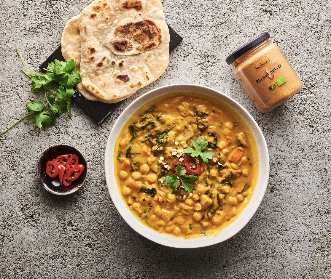 A bowl of chickpea and vegetable curry garnished with cilantro and red chili slices, served with flatbread, a jar of peanut butter, and a small bowl of sliced red chili peppers on a textured gray surface.