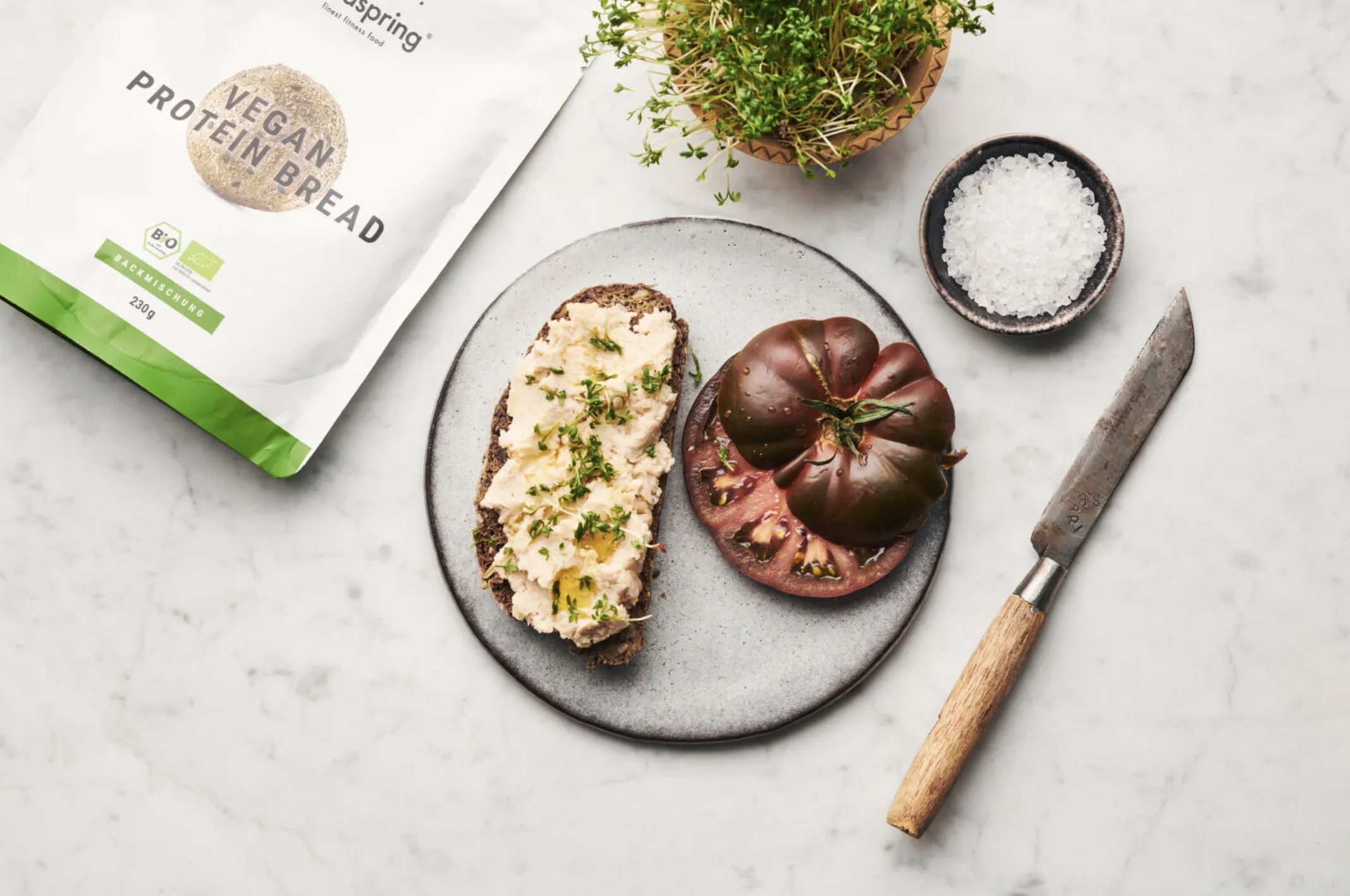 A plate with a slice of vegan protein bread topped with a spread and garnished with herbs, a slice of dark heirloom tomato, and a whole heirloom tomato on top of the tomato slice. There is a small bowl of coarse salt, a knife with a wooden handle, a 