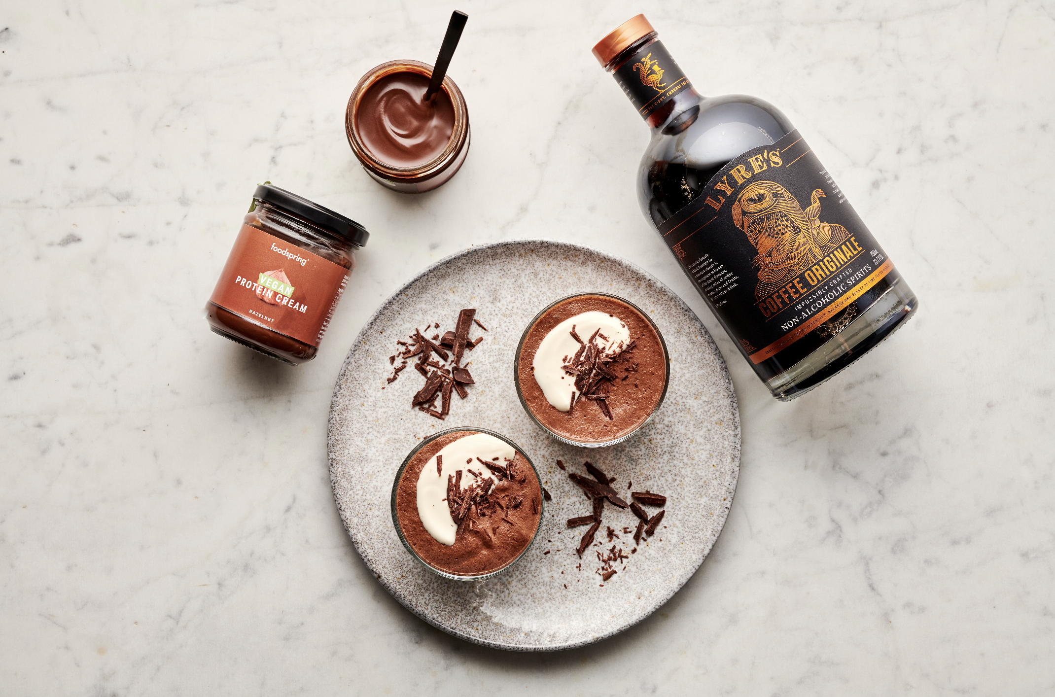 Two glasses of chocolate mousse topped with whipped cream and chocolate shavings on a round gray plate, with a jar of vegan protein cream, jar of chocolate spread, and a bottle of LAYRE'S Coffee Original non-alcoholic spirits on a white marble surfac