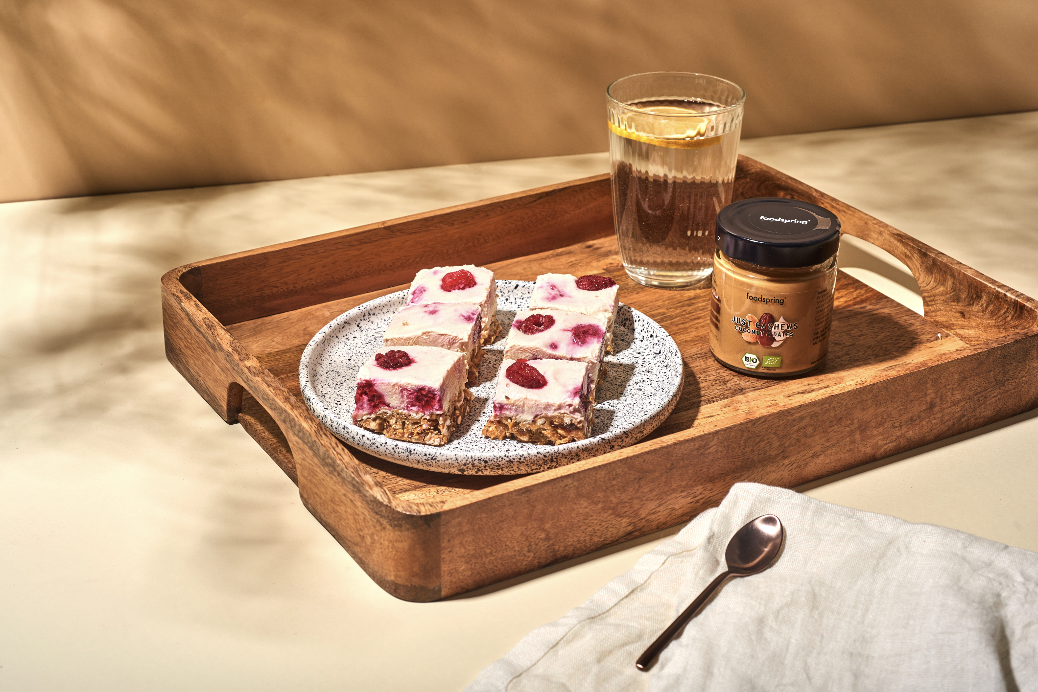 A wooden tray holding a glass of water, a jar of coconut and date spread, and a plate of berry-flavored dessert with a creamy topping and raspberries.