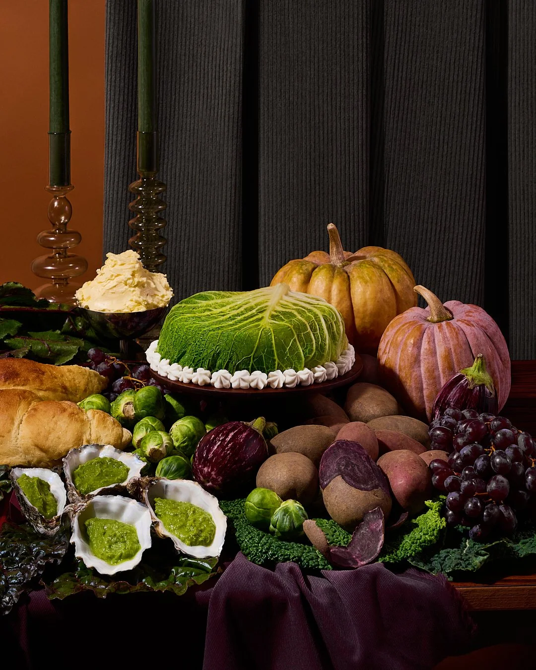 Autumn harvest display with pumpkins, brussels sprouts, grapes, purple eggplant, apples, and a cabbage-shaped cake.