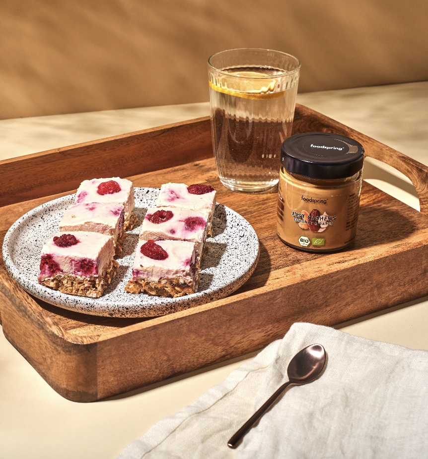 A wooden tray with a glass of water with lemon, a jar of almond butter, and a plate of rectangular berry cheesecake bites on a speckled white plate, with a small spoon on a white napkin in front.