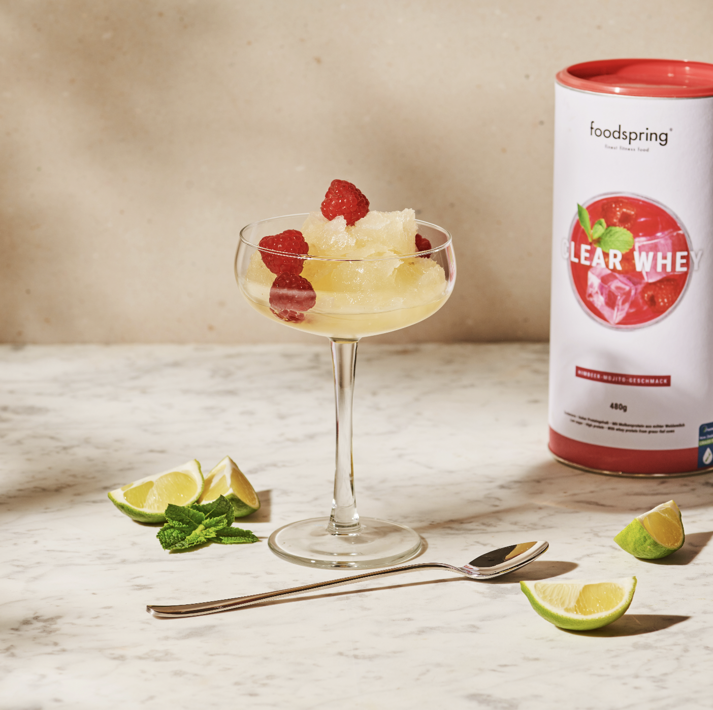 A cocktail glass filled with a yellowish frozen drink garnished with raspberries. Nearby are lime wedges, mint leaves, and a silver spoon on a white marble surface. In the background, to the right, is a can of foodspring Clear Whey powder.