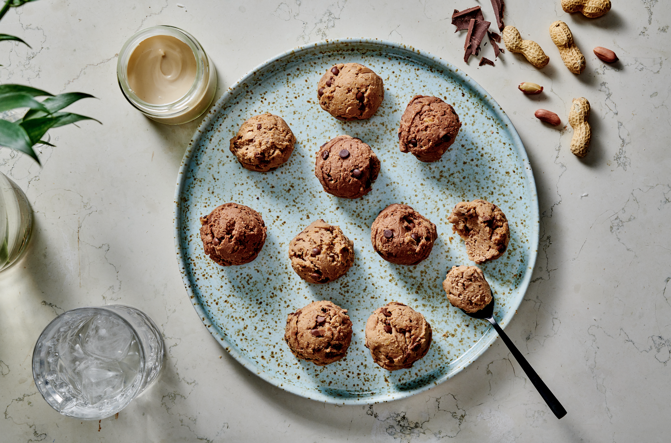 A blue speckled plate holding multiple chocolate chip cookie dough balls, some with chocolate chips, with a spoon and a piece removed. Around the plate are a glass of water, a small jar of creamy spread, and scattered nuts and chocolate pieces on a w