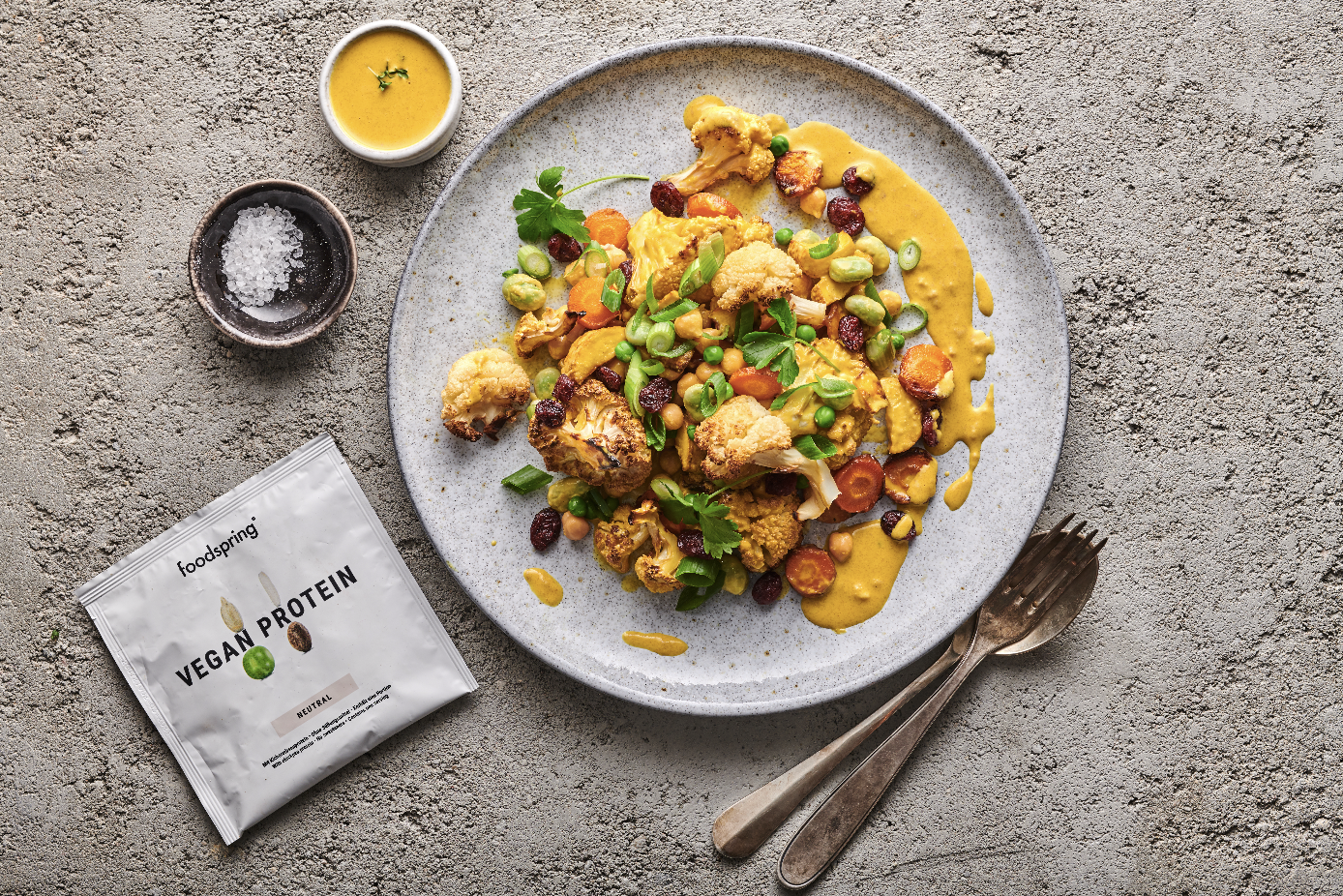 Vegan protein dish on a gray speckled plate with cauliflower, carrots, peas, and chopped green onions, drizzled with yellow sauce. Two small bowls of sauce or condiments and a packet labeled "Vegan Protein" are next to the plate, with a fork and spoo