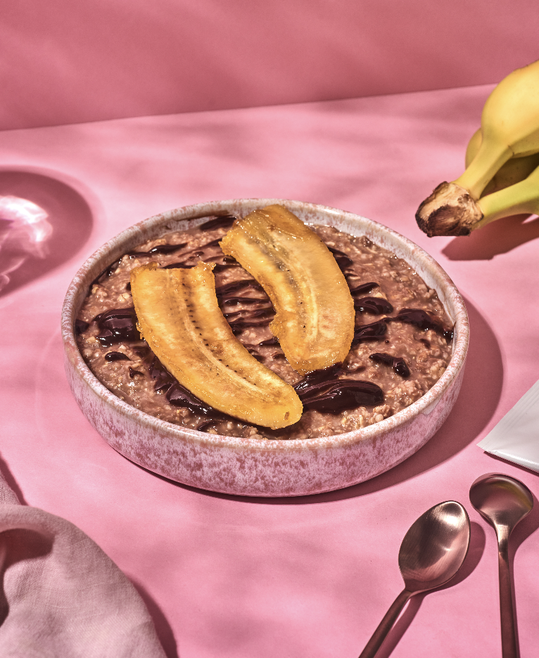 Bowl of oatmeal topped with sliced bananas and chocolate drizzle on a pink table with gold spoons nearby.