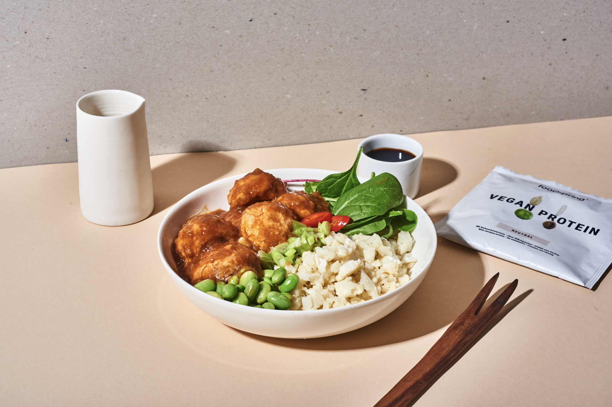 A bowl of vegan food containing tofu, edamame, greens, cherry tomatoes, and pickled onions, accompanied by soy sauce, a white ceramic pitcher, and a packet of vegan protein, on a light-colored surface with a neutral background.