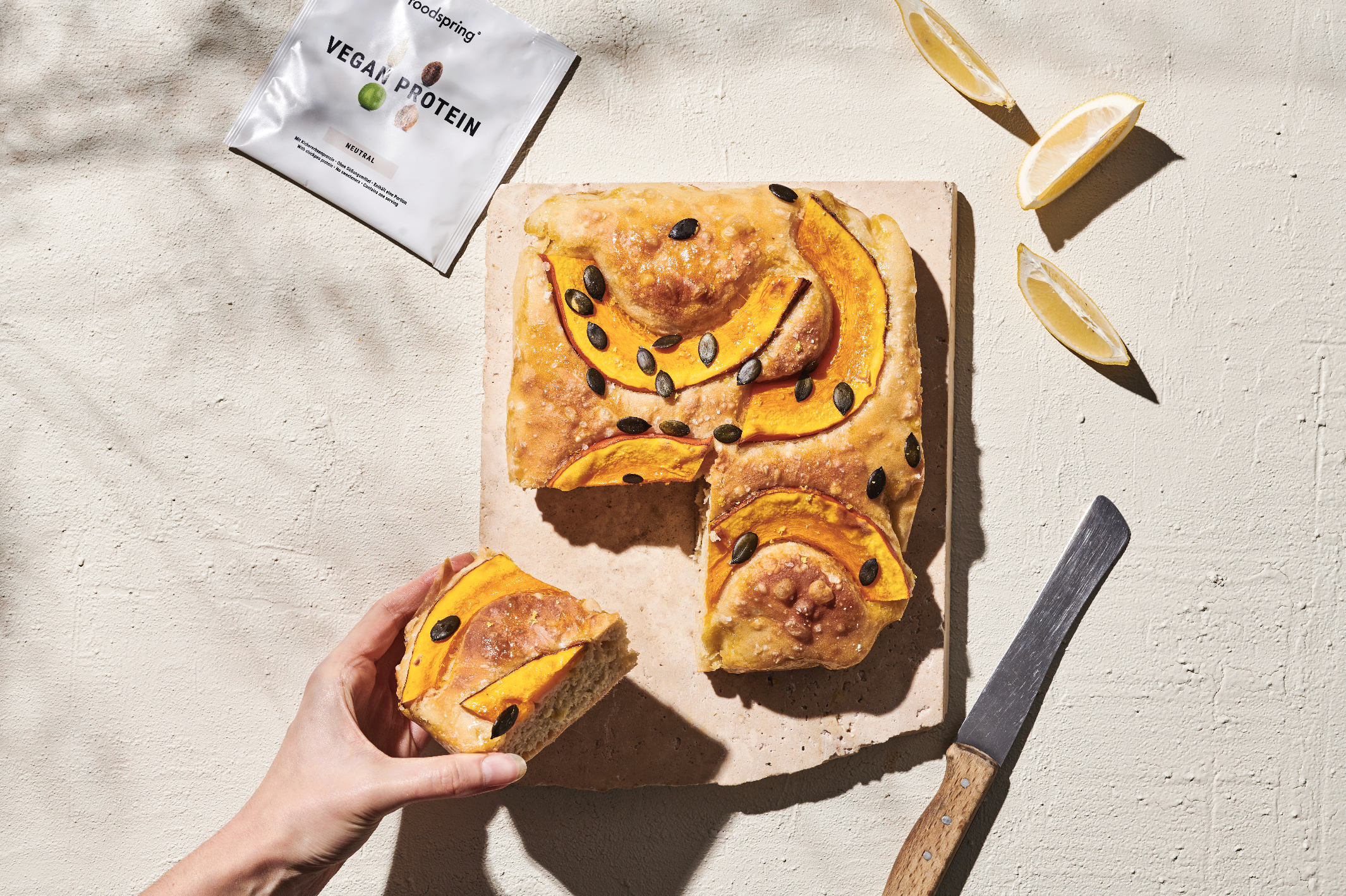 A hand holding a slice of flatbread pizza with pumpkin slices and pumpkin seeds on top. The pizza is on a pink marble cutting board. Whole lemon wedges and a lemon slice are nearby. A silver knife with a wooden handle is on the right. A packet of veg