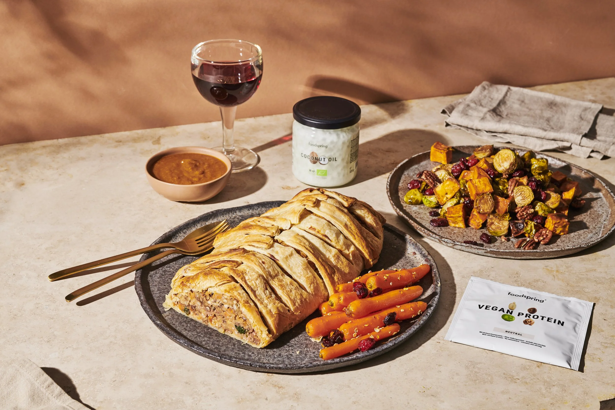 A dinner setup with a glass of red wine, a bowl of sauce, a jar of coconut oil, two plates with roasted vegetables, a vegan protein roll with carrots and berries, and a packet of vegan protein powder on a light-colored table.
