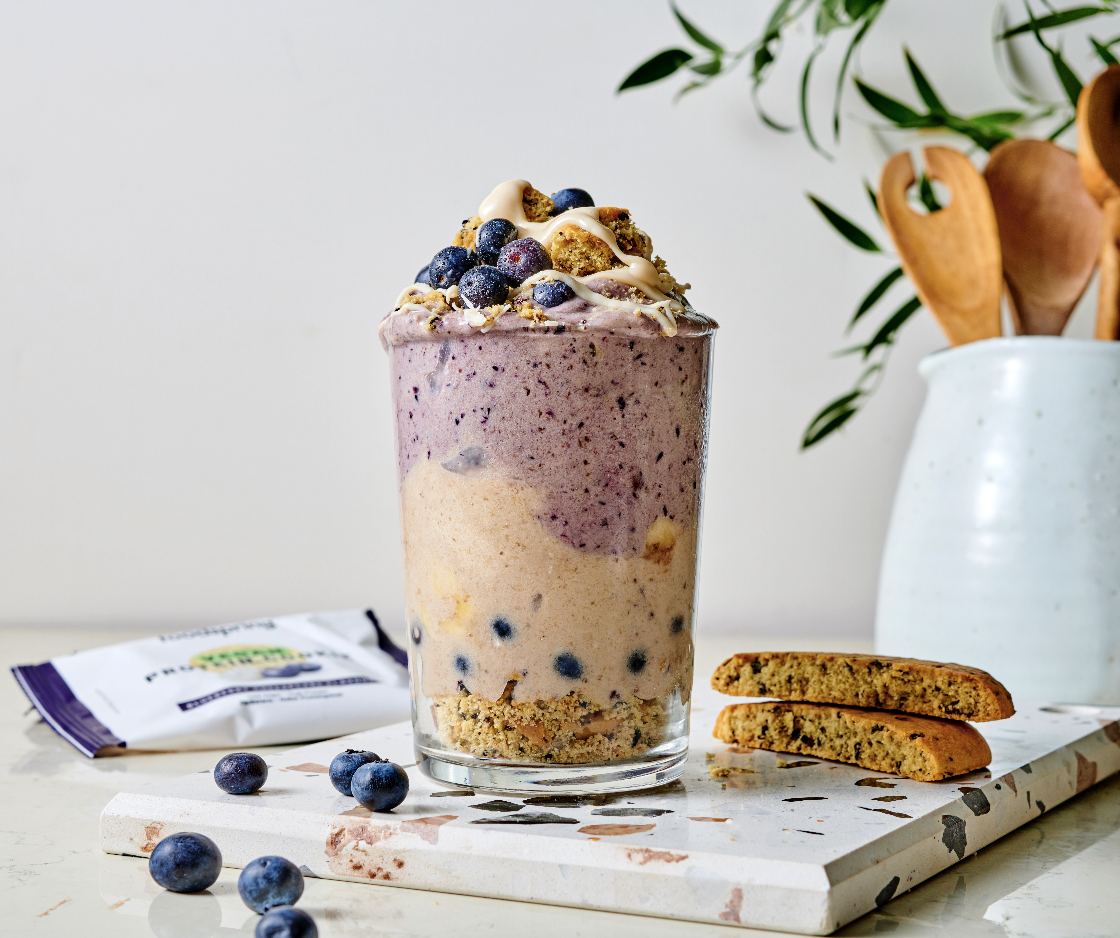 A layered blueberry smoothie in a clear glass topped with blueberries, crumbled cookies, and white sauce, on a white marble surface with scattered blueberries, a couple of cookies, and a white ceramic vase with orange flowers and green leaves in the background.