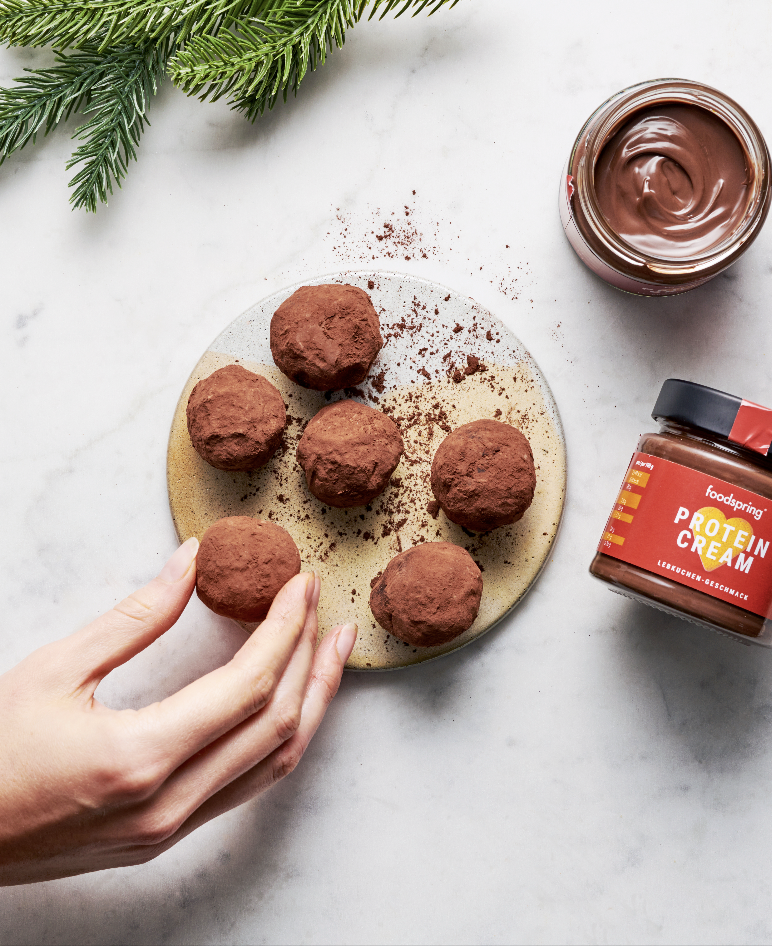 Chocolate protein energy bites on a plate, with a jar of chocolate protein cream and a jar of cocoa powder, with green pine branches in the top left corner on a white surface.