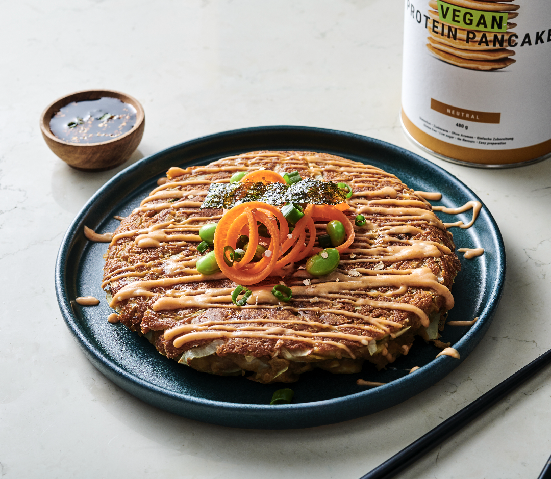 Vegan protein pancake topped with sliced carrots, edamame, and seaweed slices, drizzled with sauce on a dark plate, with a container labeled 'Vegan Protein Pancake' in the background.
