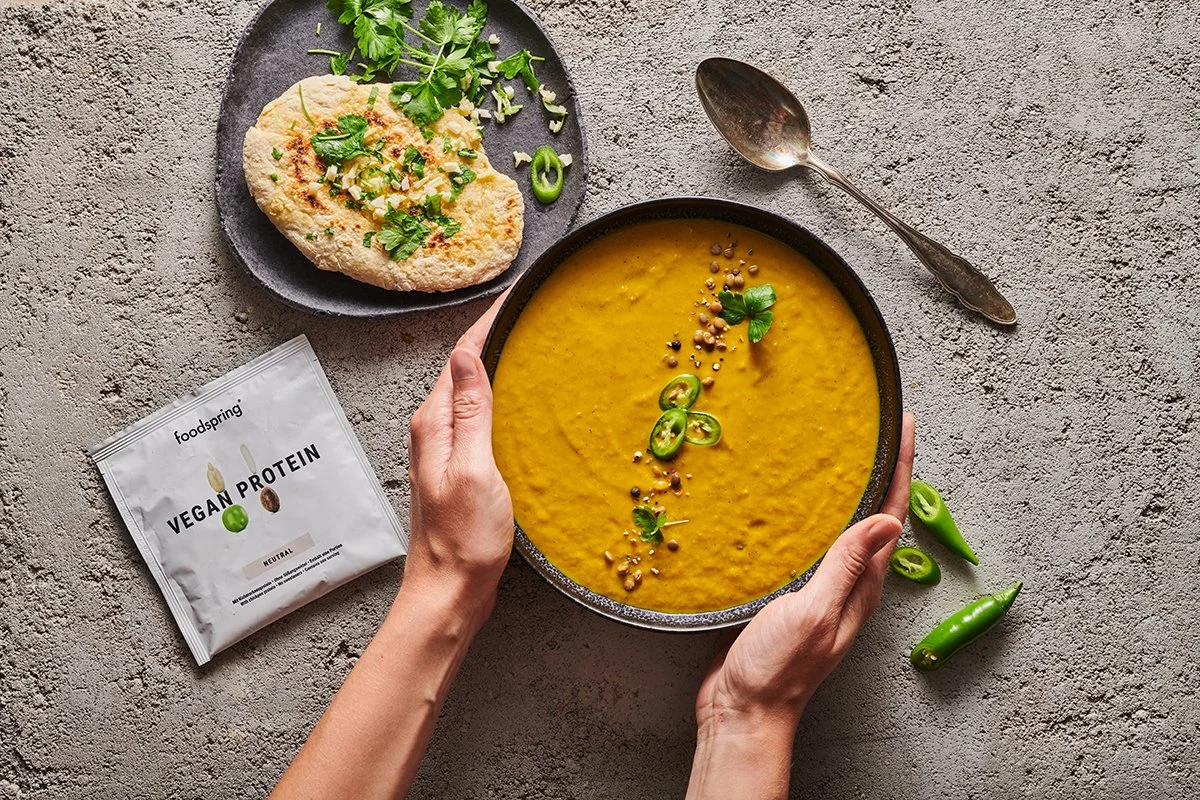 A bowl of yellow vegan soup garnished with green chili slices and herbs, with a side of flatbread topped with chopped herbs, parsley, and green onions, on a textured gray surface.