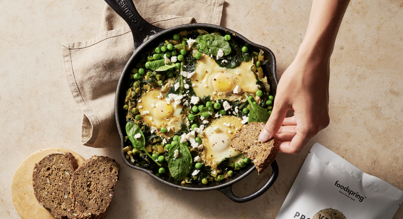 Skillet with eggs, spinach, green peas, and crumbled cheese, with slices of multigrain bread on a plate and a hand holding a piece of bread over the skillet.