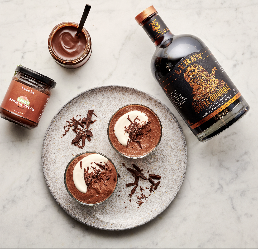 Chocolate mousse dessert in two glasses garnished with whipped cream and chocolate shavings on a gray plate, with a jar of vegan protein cream, a jar of chocolate spread, and a bottle of coffee-flavored liquor on a white marble surface.