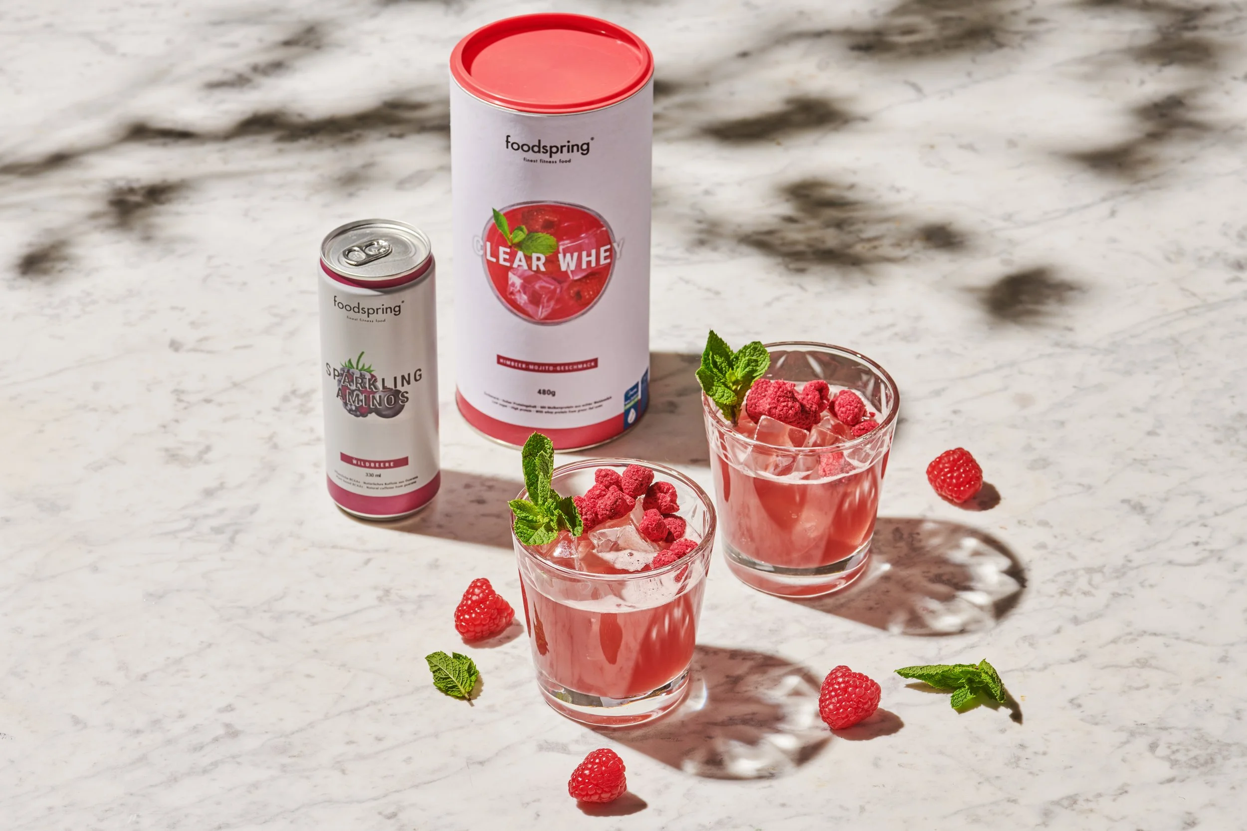 Two glasses of raspberry-flavored beverage with raspberries and mint leaves on a white marble surface, with canned raspberry-flavored drink mixes and a container in the background.