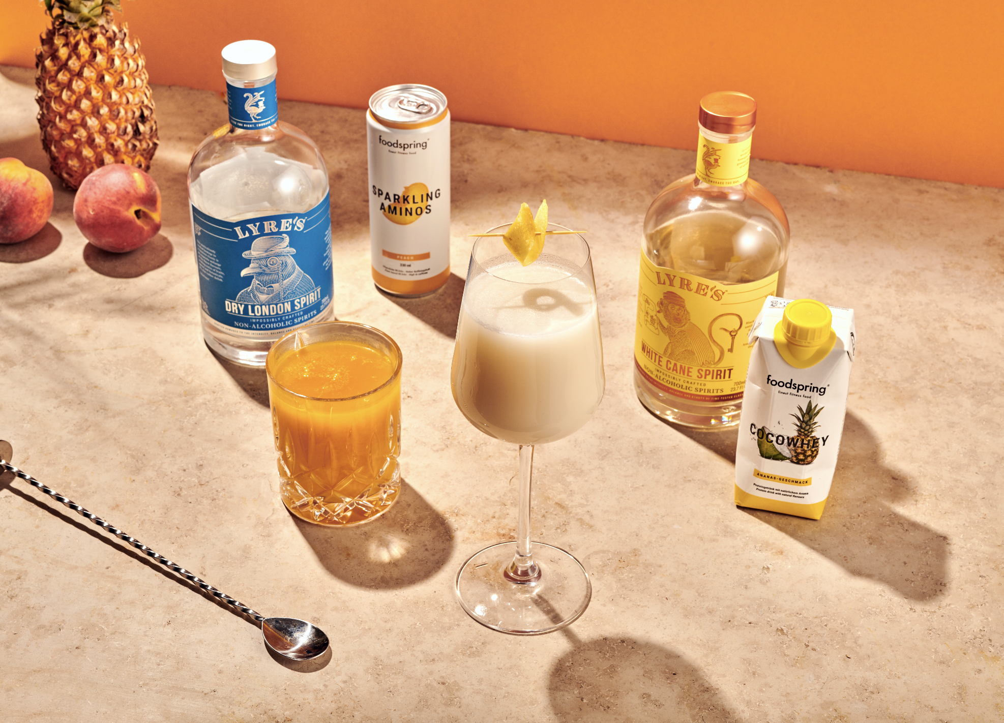 An assortment of beverages on a countertop, including two bottles of flavored spirits, a can of sparkling amino, a carton of coconut water, a glass of orange juice, a cocktail with a lemon garnish, and a pineapple, with two peaches nearby.