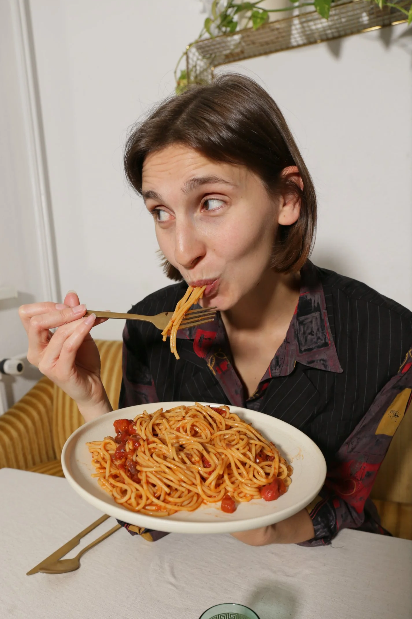 A woman with brown hair, wearing a black shirt, holding a plate of spaghetti with tomato sauce, with spaghetti in her mouth using a fork.