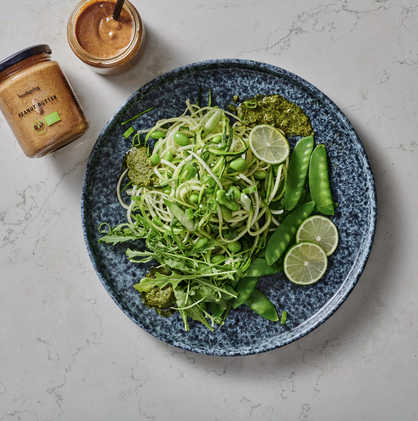 A blue bowl filled with spiralized zucchini, snap peas, broccoli, sliced limes, and pesto sauce, with jars of peanut butter and a spoon of peanut butter on the side on a white marble surface.