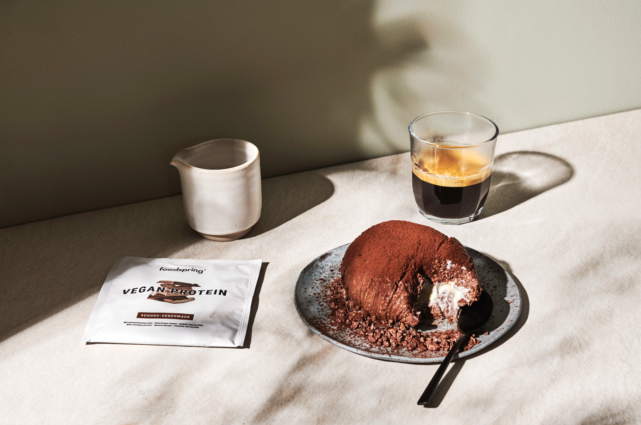 Chocolate dessert on a plate with a spoon, a glass of black coffee with ice, a white pitcher, a vegan protein pack labeled "Sweet Taste," and a beige background with shadows.