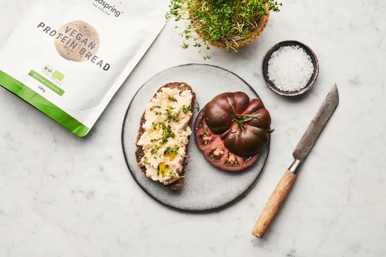 Open package of vegan protein bread next to a plate with sliced tomato and hummus on toast, a small bowl of sea salt, a knife, and a potted plant on a white marble surface.