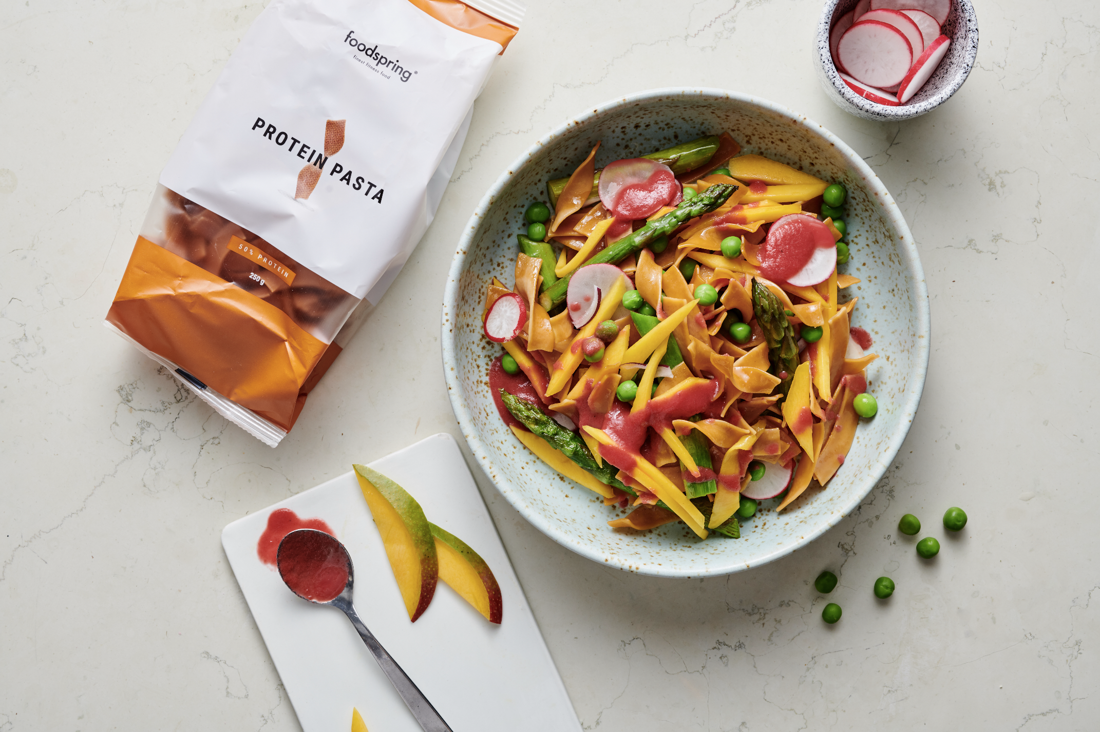 Bowls of pasta salad with radishes, green peas, and asparagus on a white surface. A bag of protein pasta from foodspring is nearby, along with a small dish of radish slices and a white plate with mango and cantaloupe slices. A spoon with red sauce is