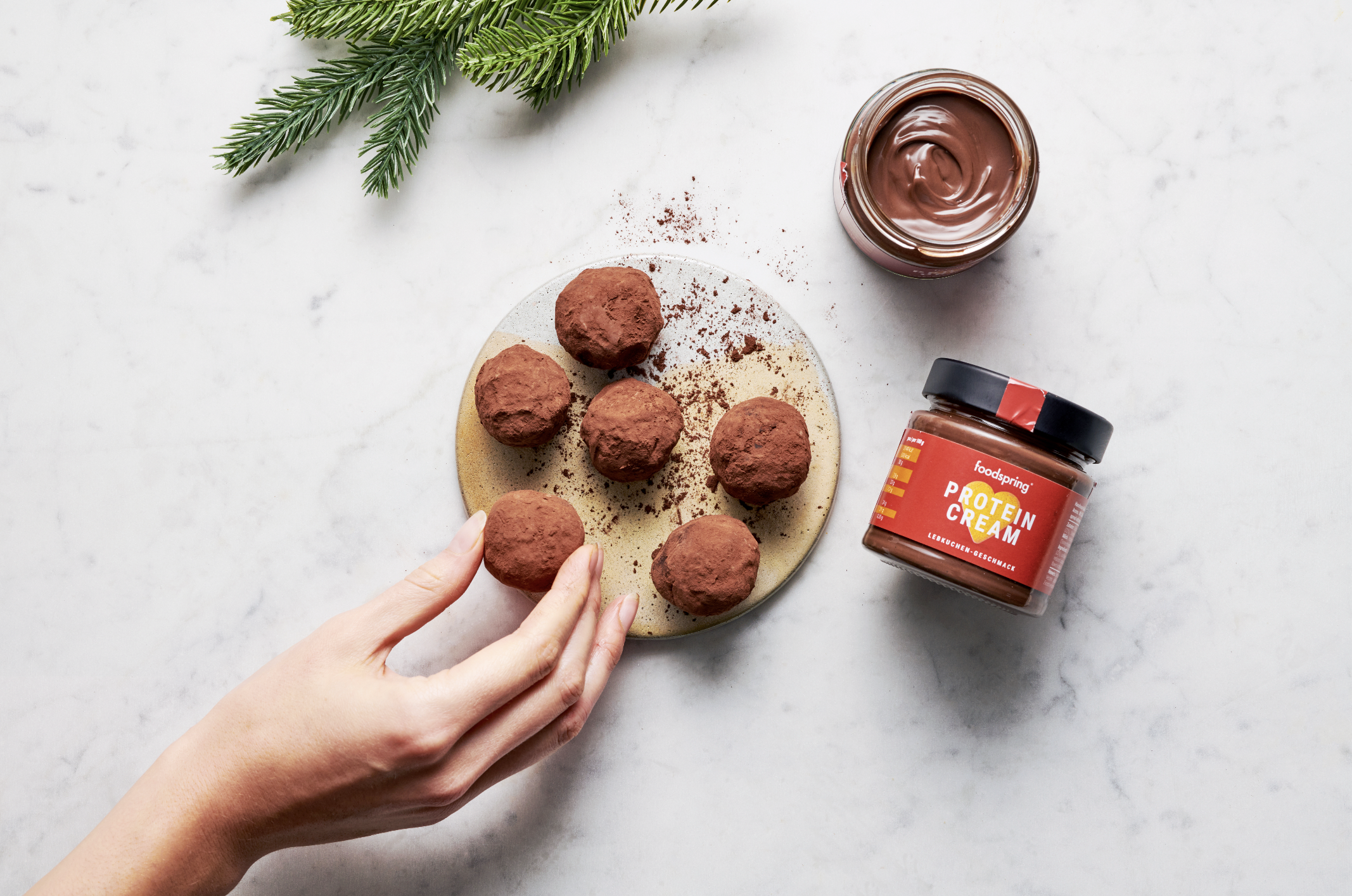 A hand placing a chocolate truffle on a beige plate, with additional truffles sprinkled with cocoa powder, a jar of chocolate hazelnut spread, a jar of protein chocolate cream, and some green pine branches on a white marble surface.