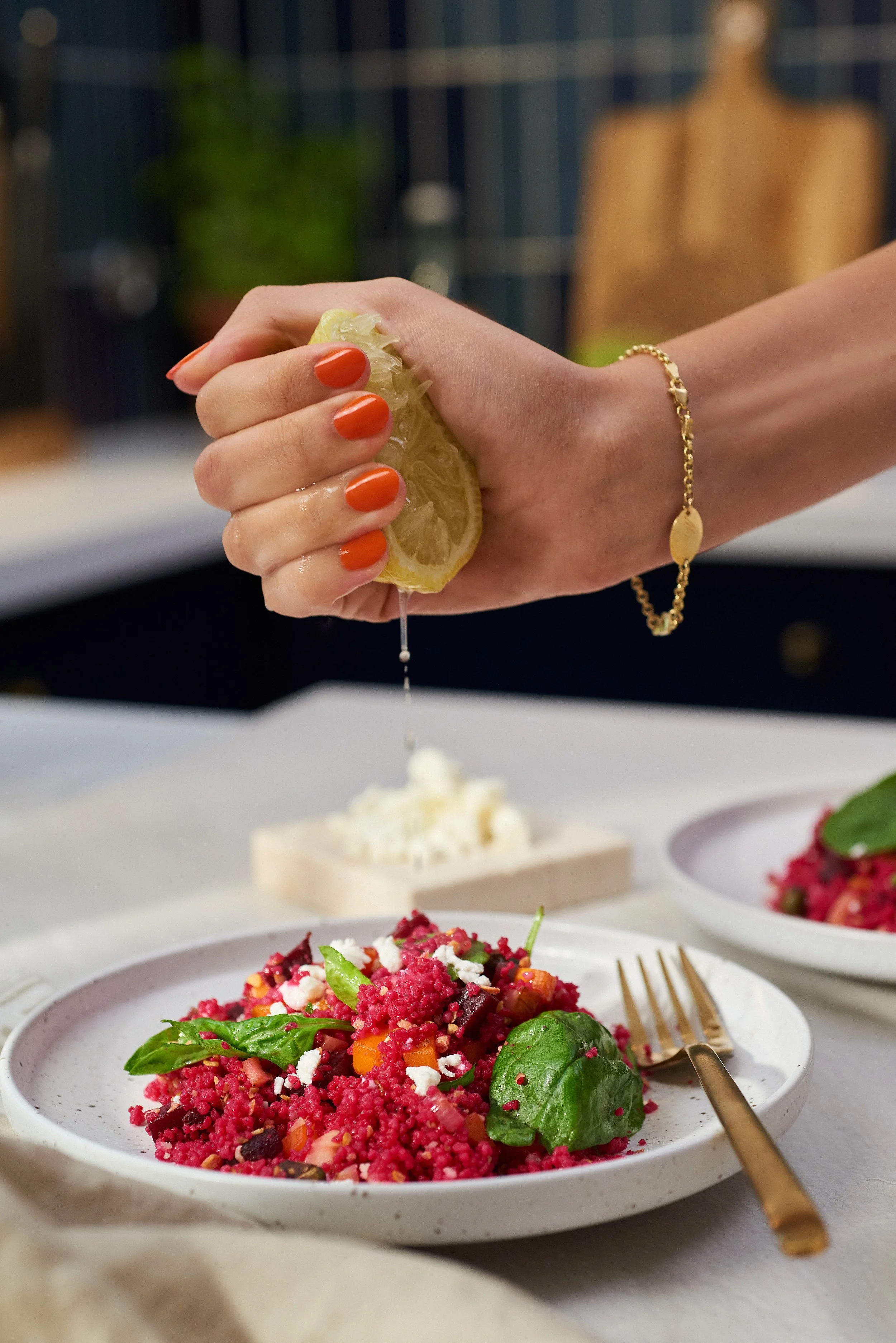 Person squeezing lemon over a colorful beet and vegetable salad on a white plate, with a gold fork and another plate of salad in the background.