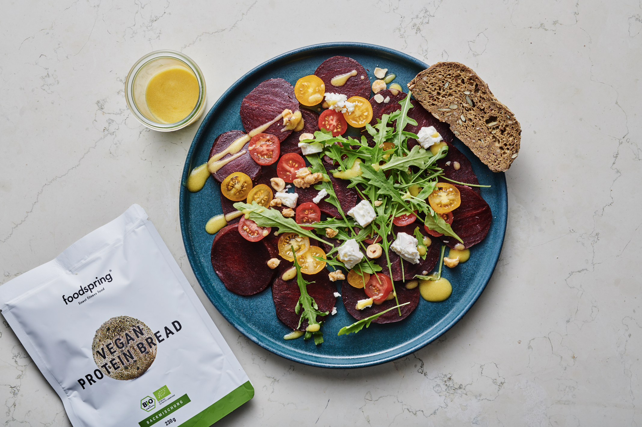 A blue plate with a salad containing cherry tomatoes, arugula, beet slices, and cheese, with a slice of multigrain bread on the edge. A small bowl of dressing, a package of vegan protein bread, and a glass of yellow dressing or juice are also on the 