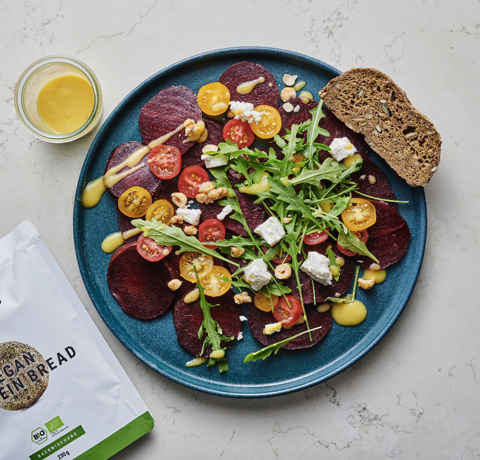 Beet and cherry tomato salad on a blue plate with arugula, crumbled cheese, chopped nuts, and a slice of whole grain bread, with a small glass of yellow dressing or juice nearby and a bag of organic rye bread on the side.