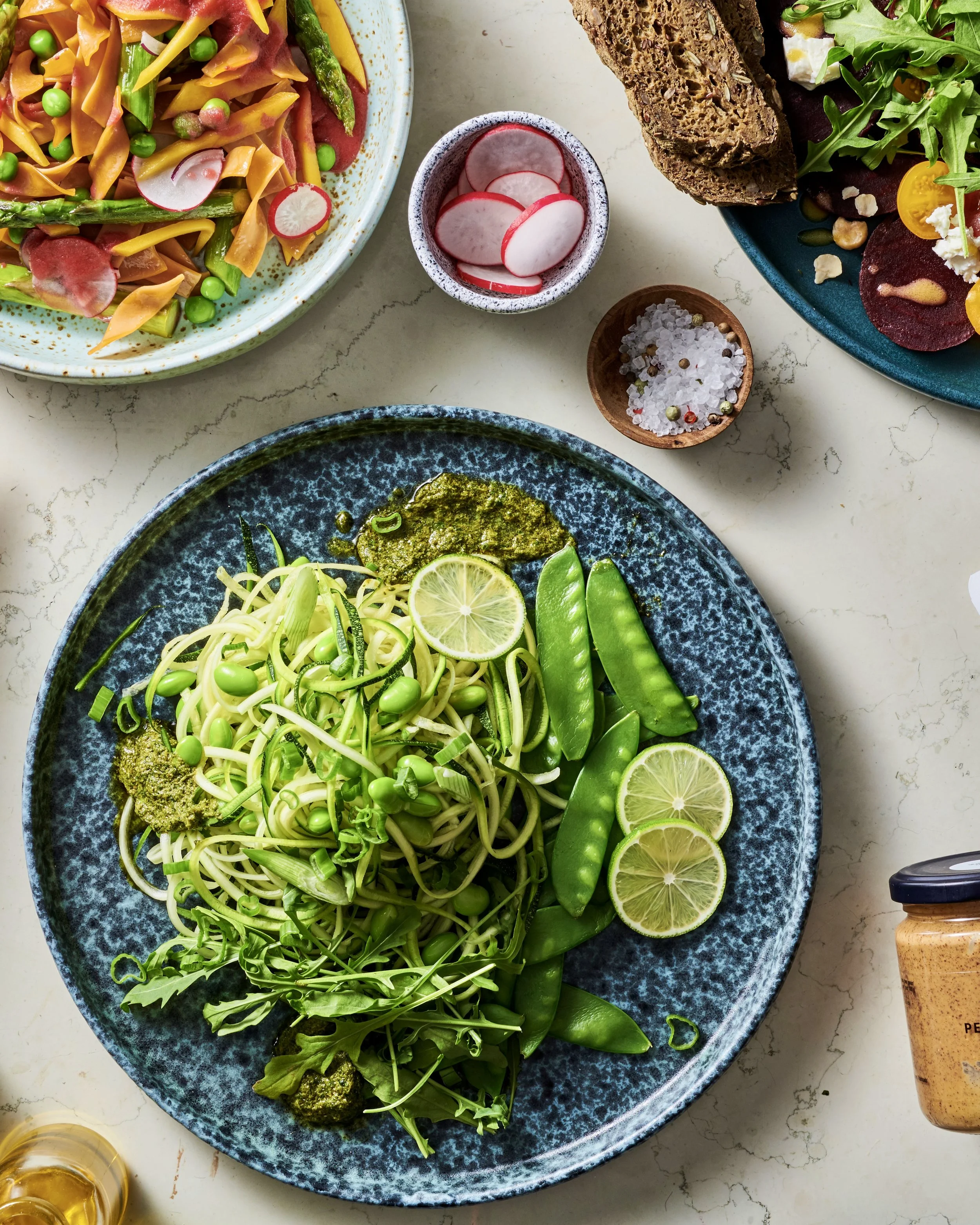 A plate of green noodles with pea pods, lime slices, pesto, and microgreens. Surrounding dishes include a salad with radishes, a plate with bread and assorted meats, and small bowls of radish slices, coarse salt, and mustard.