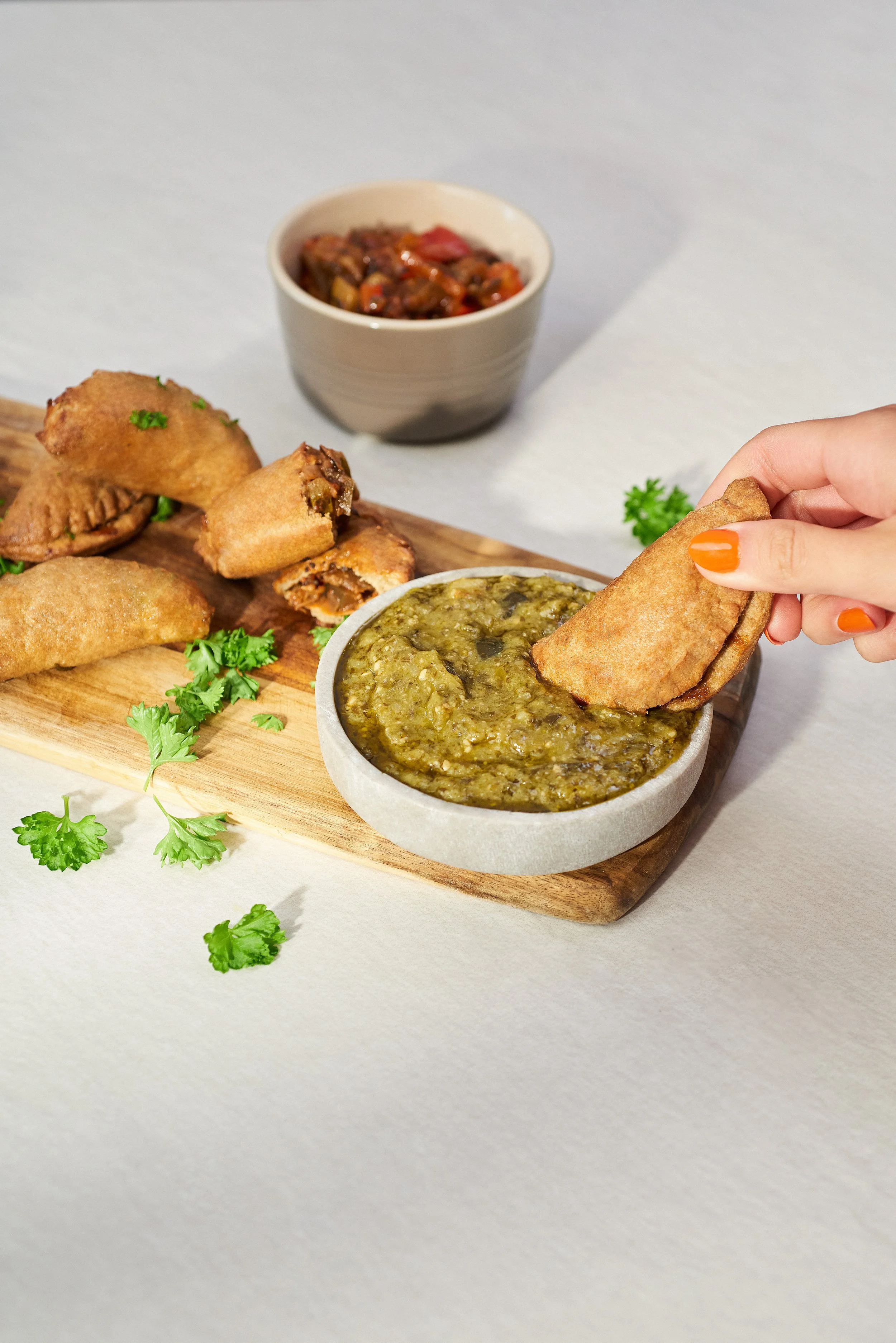 Person dipping a samosa into a bowl of green chutney, with fried samosas and a bowl of salsa on a wooden board.