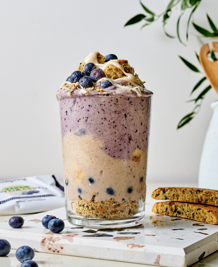 A layered blueberry dessert in a clear glass, topped with blueberries, white chocolate chips, and cookie crumbles, with cookies and a leafy plant in the background.