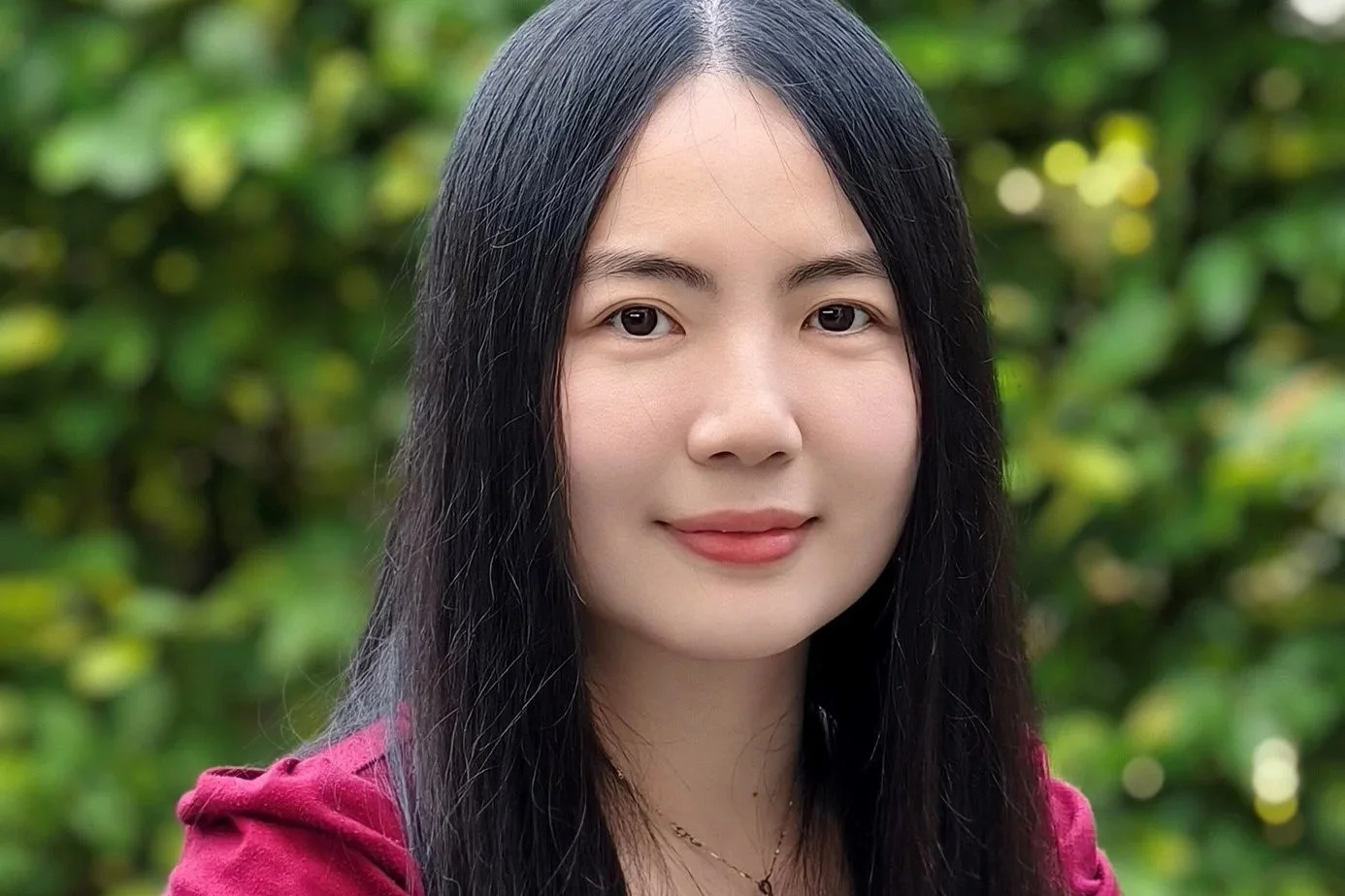A young woman with long black hair and fair skin outdoors, wearing a burgundy top, with a blurred green leafy background.