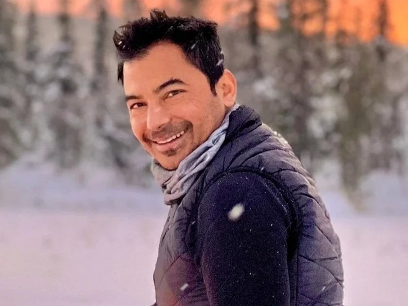 A smiling man outdoors in winter, wearing a dark jacket and scarf, with snow and trees in the background.