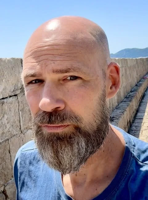 Close-up of a man with a beard and shaved head outdoors on a sunny day, with a stone wall and distant mountains in the background.