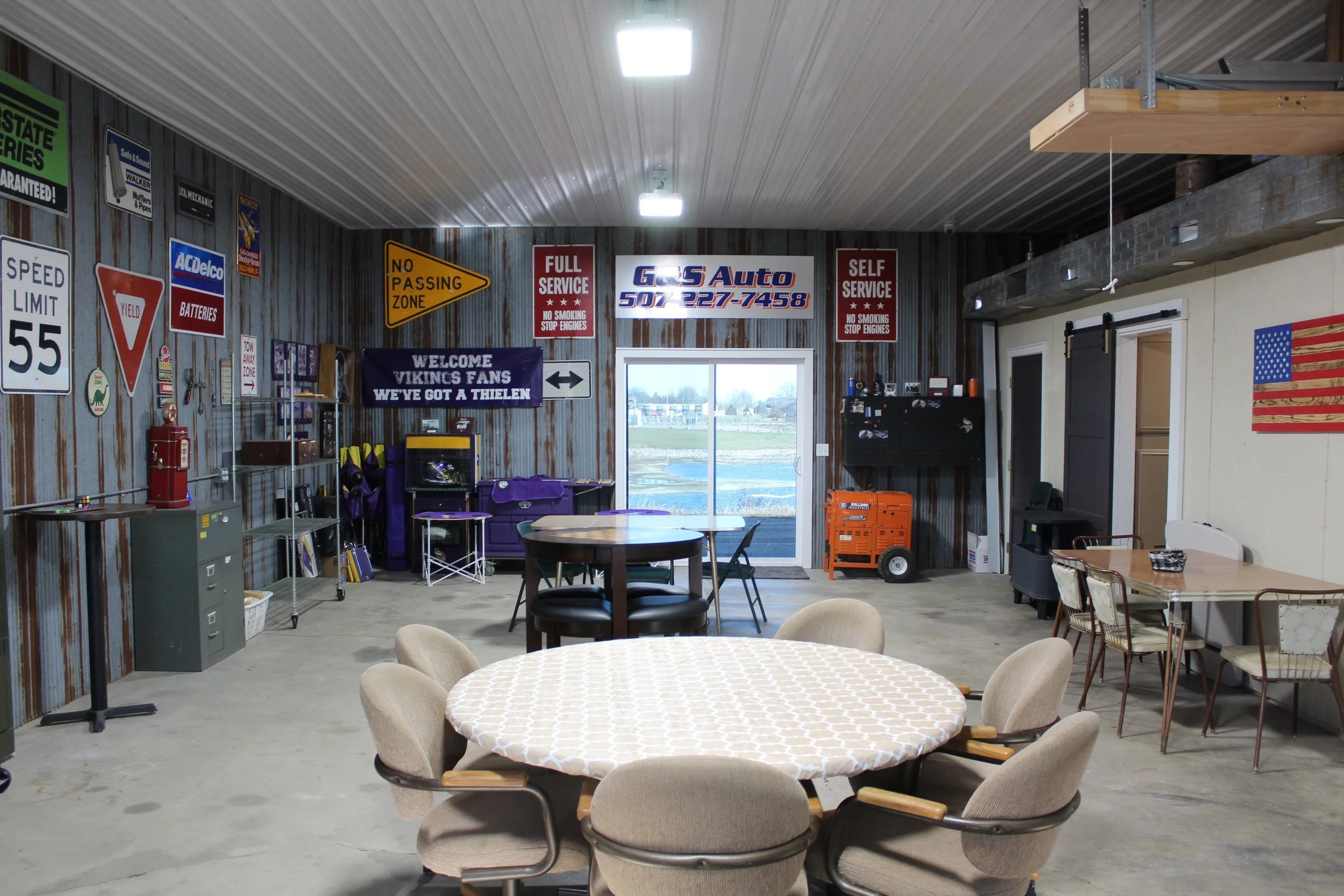 Indoor garage with tables, chairs, and vintage signs on the wall, including traffic signs, advertisements, and welcome banners.